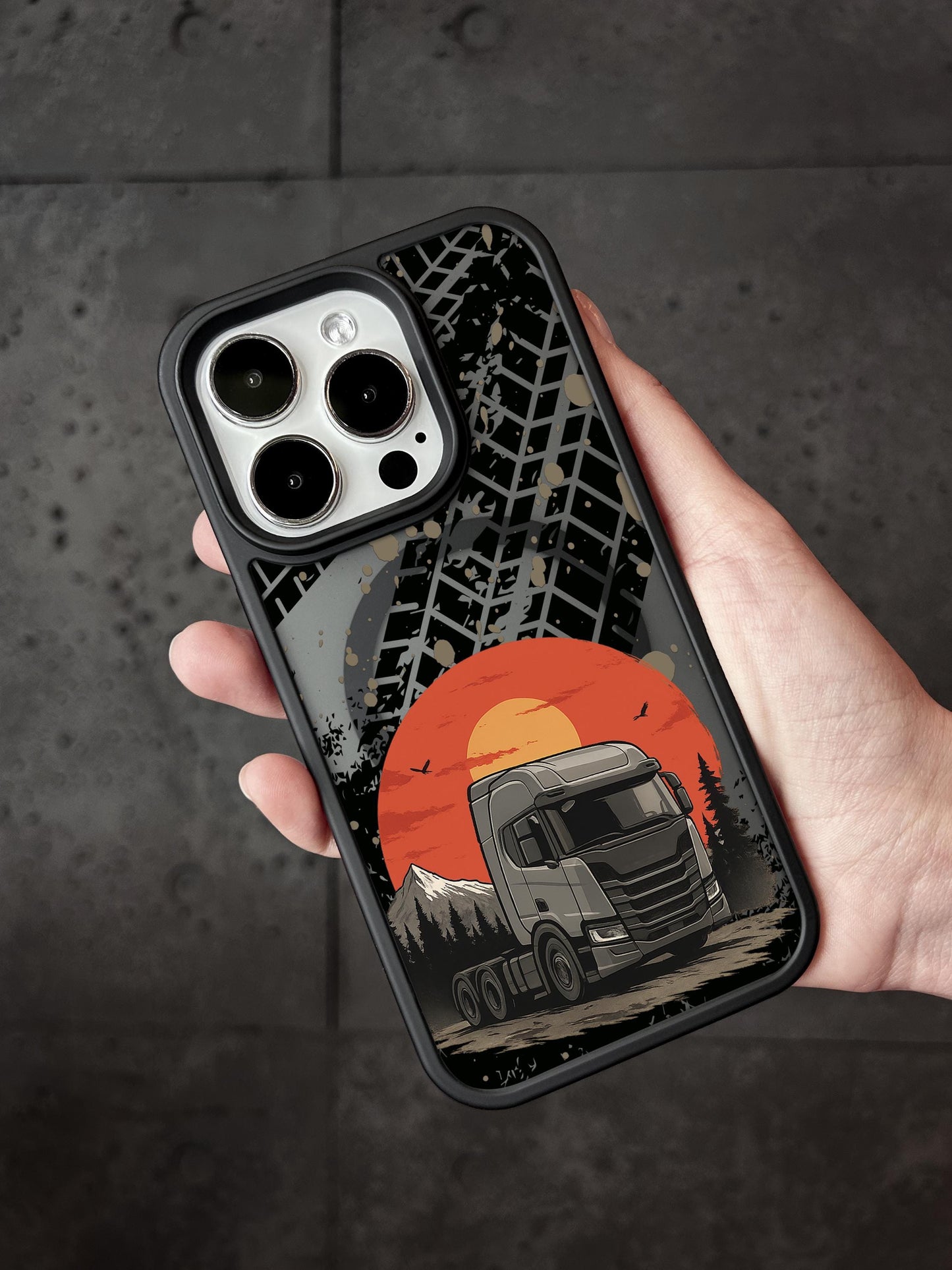 A person&#39;s hand holding a phone case with a design depicting a truck on a road against a sunset background.