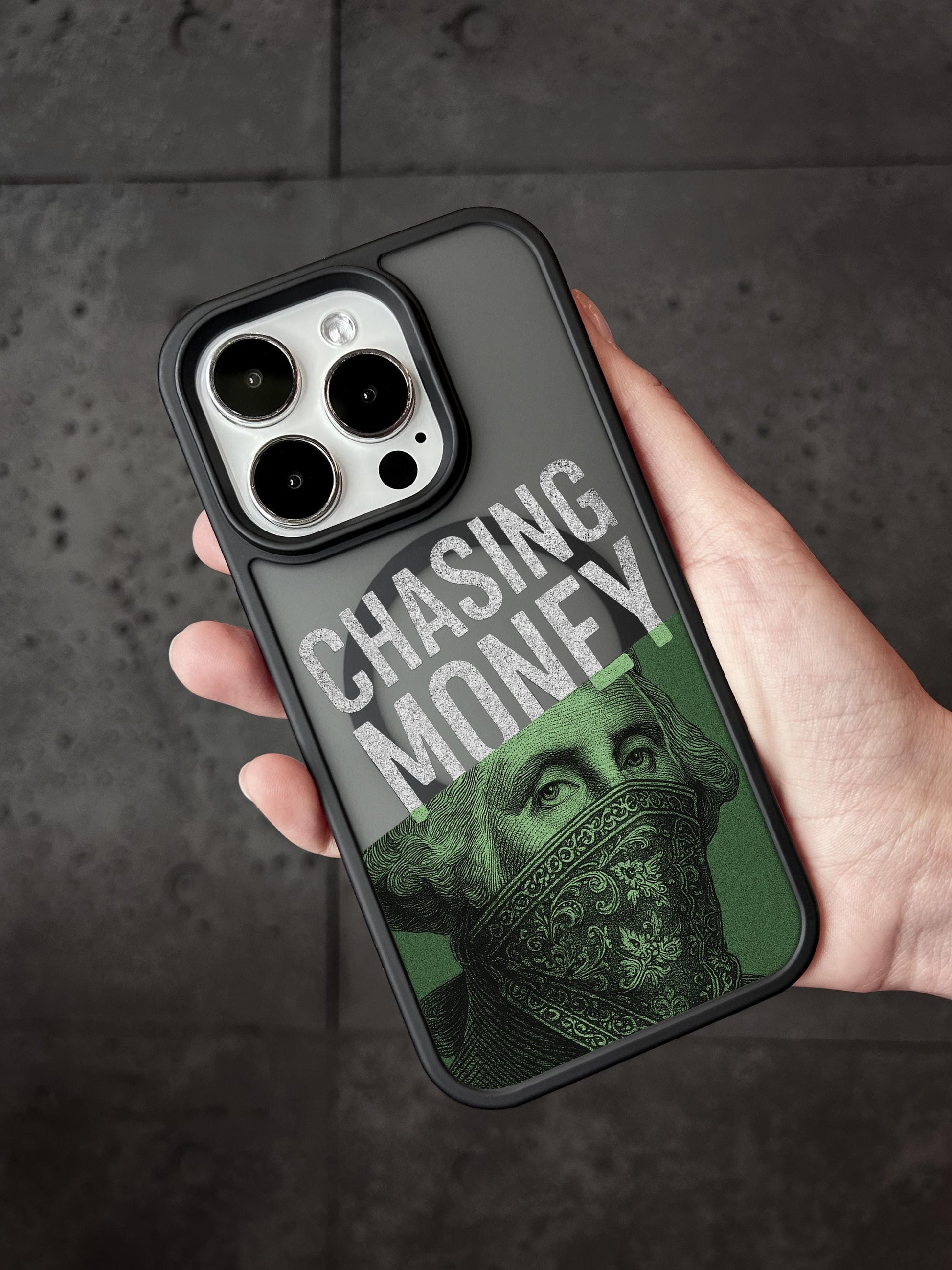 A person&#39;s hand holding a phone case with a design featuring a face wearing a bandana and the text &quot;CHASING MONEY&quot; printed on it.