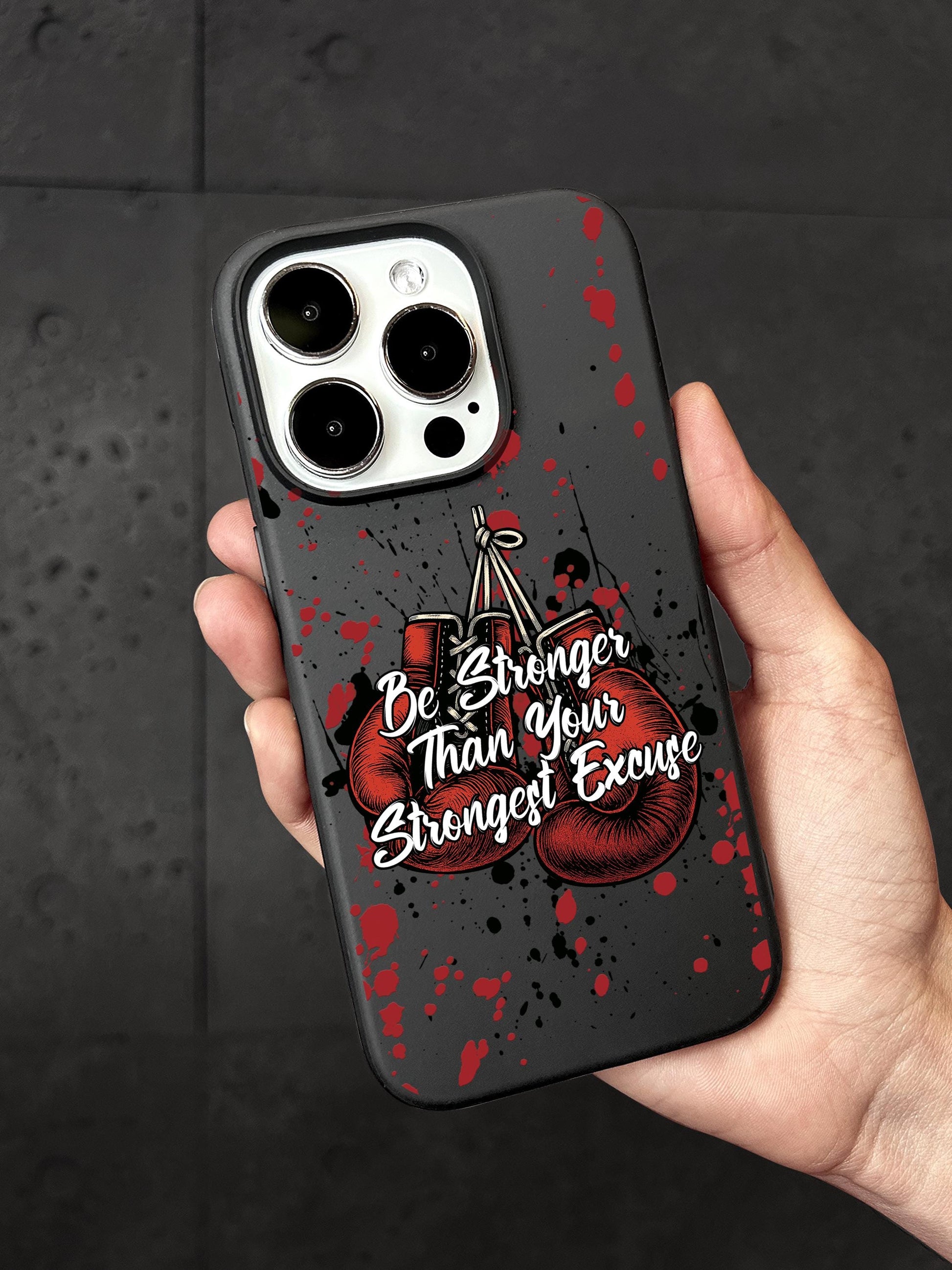 A person&#39;s hand holding a phone case with a design featuring boxing gloves and the text &quot;Be Stronger Than Your Strongest Excuse&quot;.