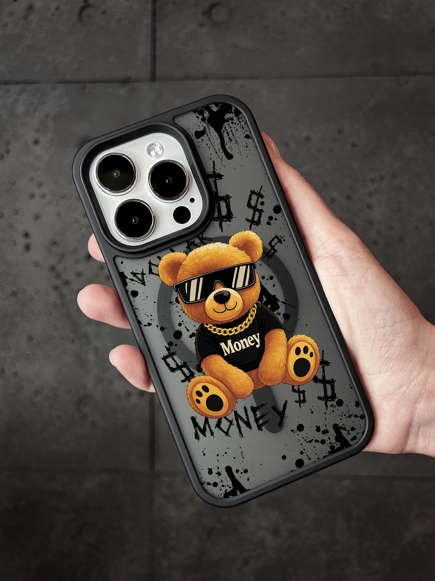 A person&#39;s hand holding a phone case featuring a teddy bear wearing sunglasses and a black shirt with the word &quot;Money&quot; printed on it.