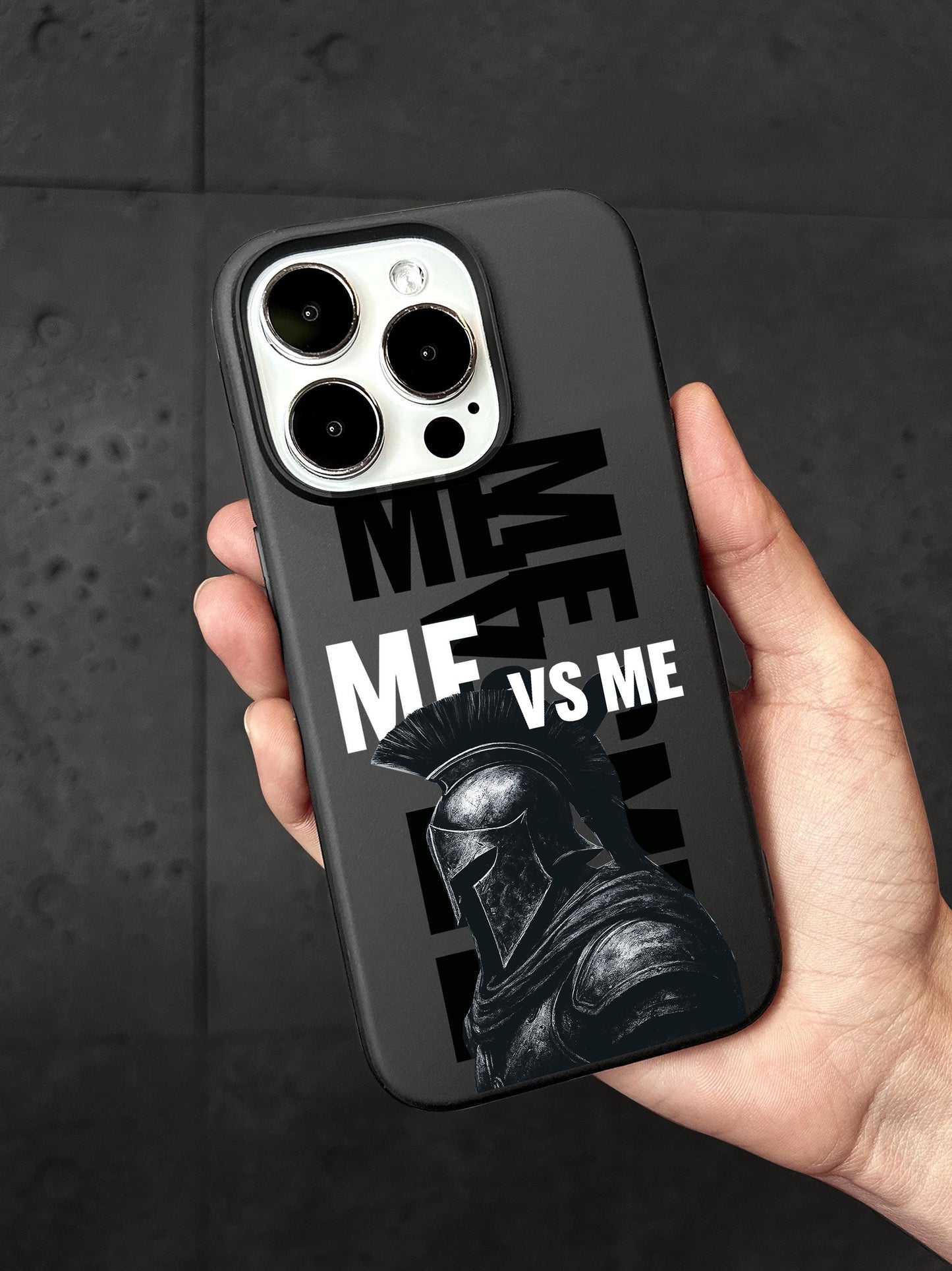 A person&#39;s hand holding a black phone case with a graphic design featuring a helmeted figure and the text &quot;ME VS ME&quot; printed on it.