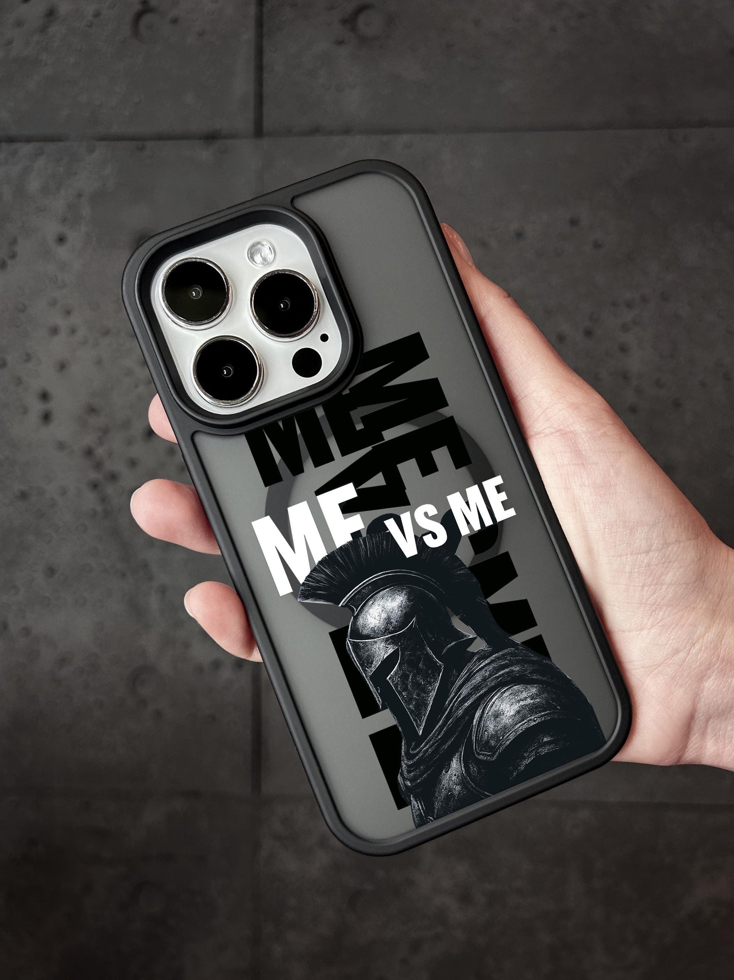 A person&#39;s hand holding a phone case with a graphic design featuring a helmeted figure and the text &quot;ME VS ME&quot; printed on it.